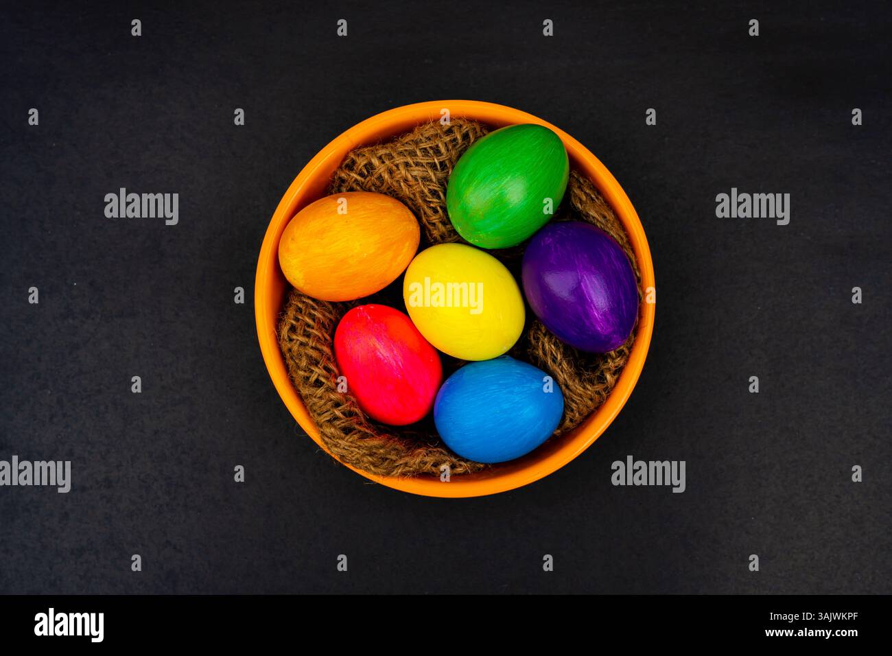 Colorful Easter Eggs in Orange Bowl on Black Background. Multicolored festive eggs displayed in a textured orange bowl with thread, styled on a dark b Stock Photo