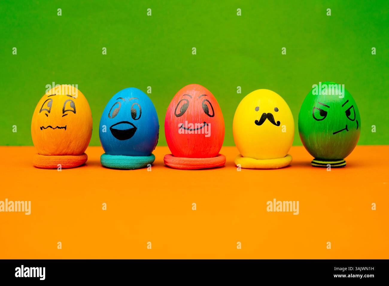 Lined Up Painted Easter Eggs With Expressive Cartoon Faces On Green and Orange Background. Playful spring-themed eggs with character designs on a bold Stock Photo