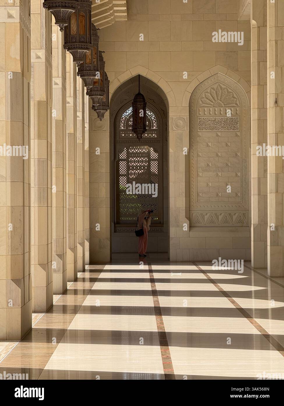 Corridor with pillars and shadows at Sultan Qaboos Grand Mosque, Muscat. A woman photographer captures a scene at the far end in daylight - Smartphone Captured Stock Image