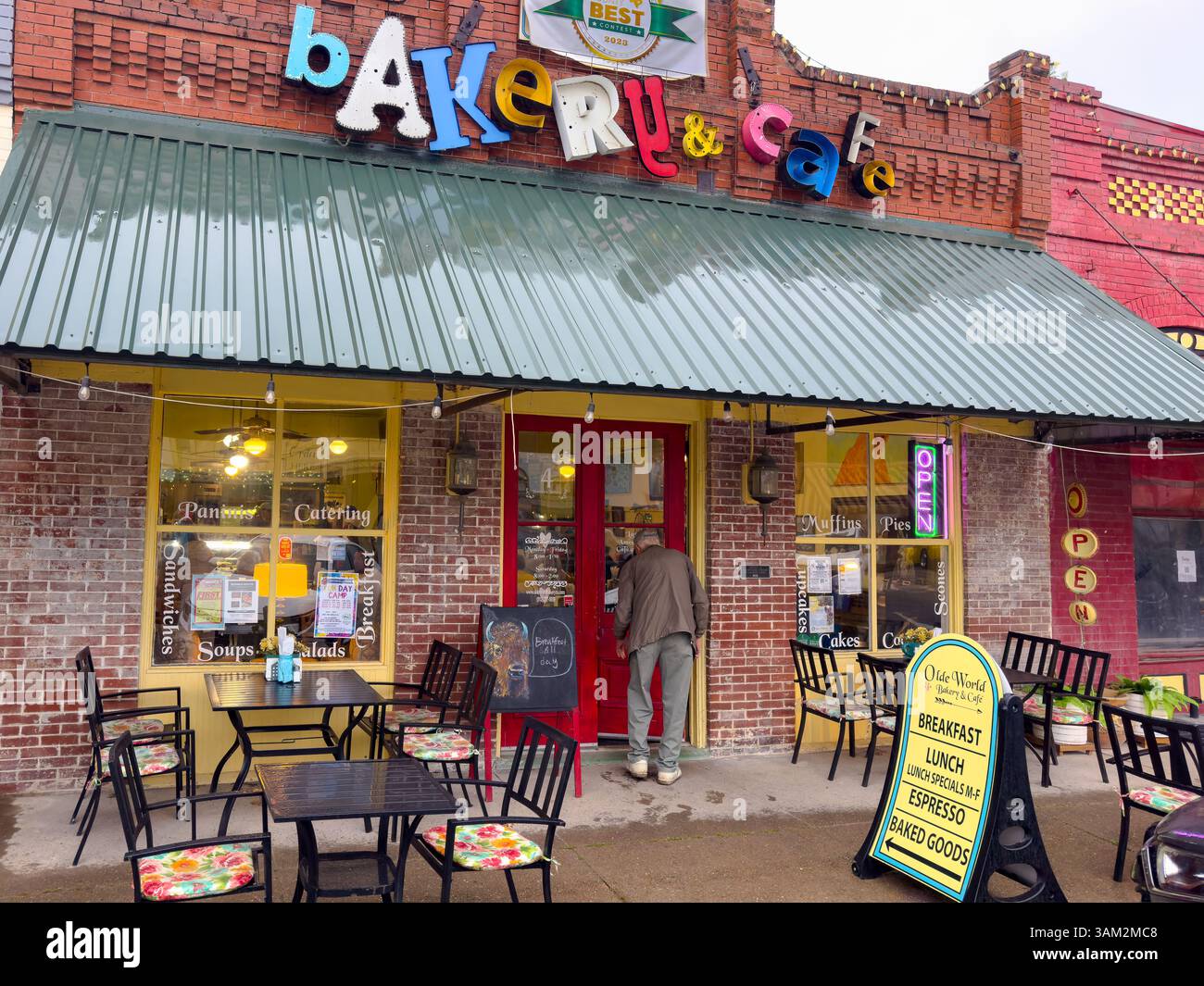 Stooped senior male walks through the red front door of Olde World Bakery & Cafe, Smithville, Bastrop County, Texas, USA. - Smartphone Captured Stock Image