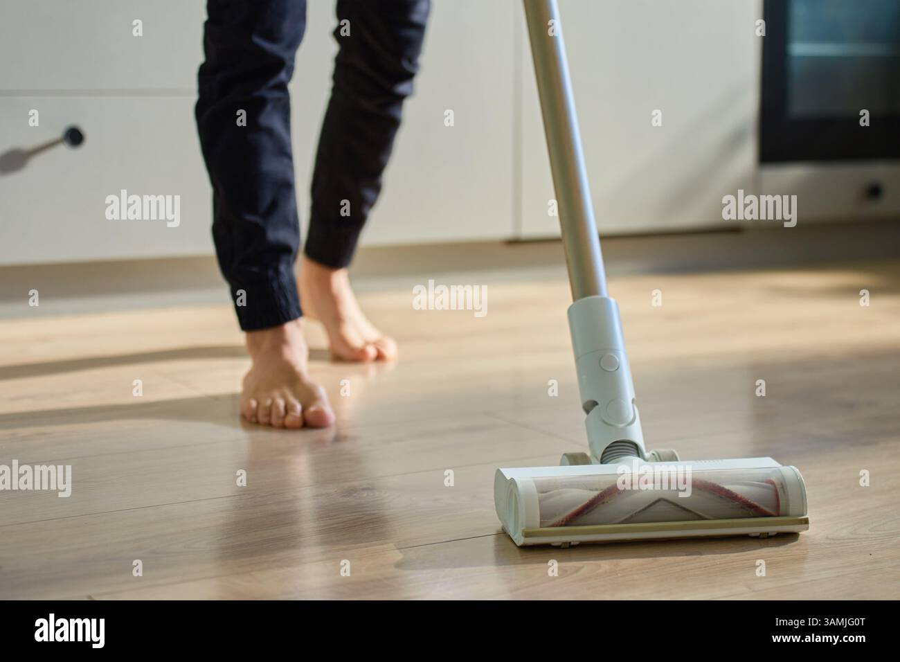 Barefoot man cleaning wooden floor in kitchen with vacuum. Person vacuuming with cordless vacuum cleaner. Daily routine and household chores Stock Photo