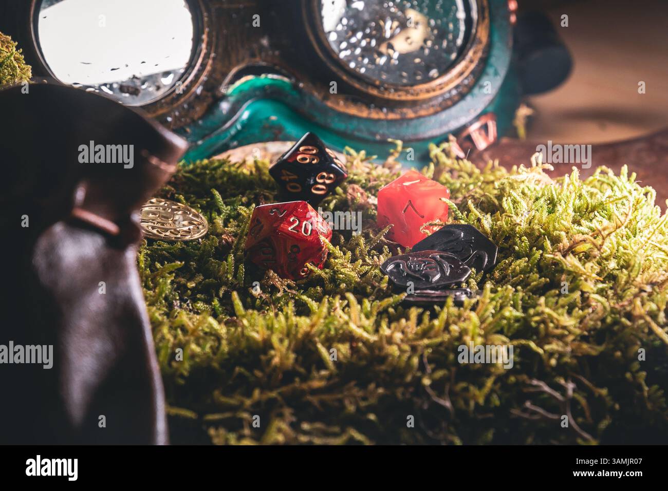 A red d20, orange d10, and black dice rest on moss near steampunk goggles and fantasy coins. Stock Photo