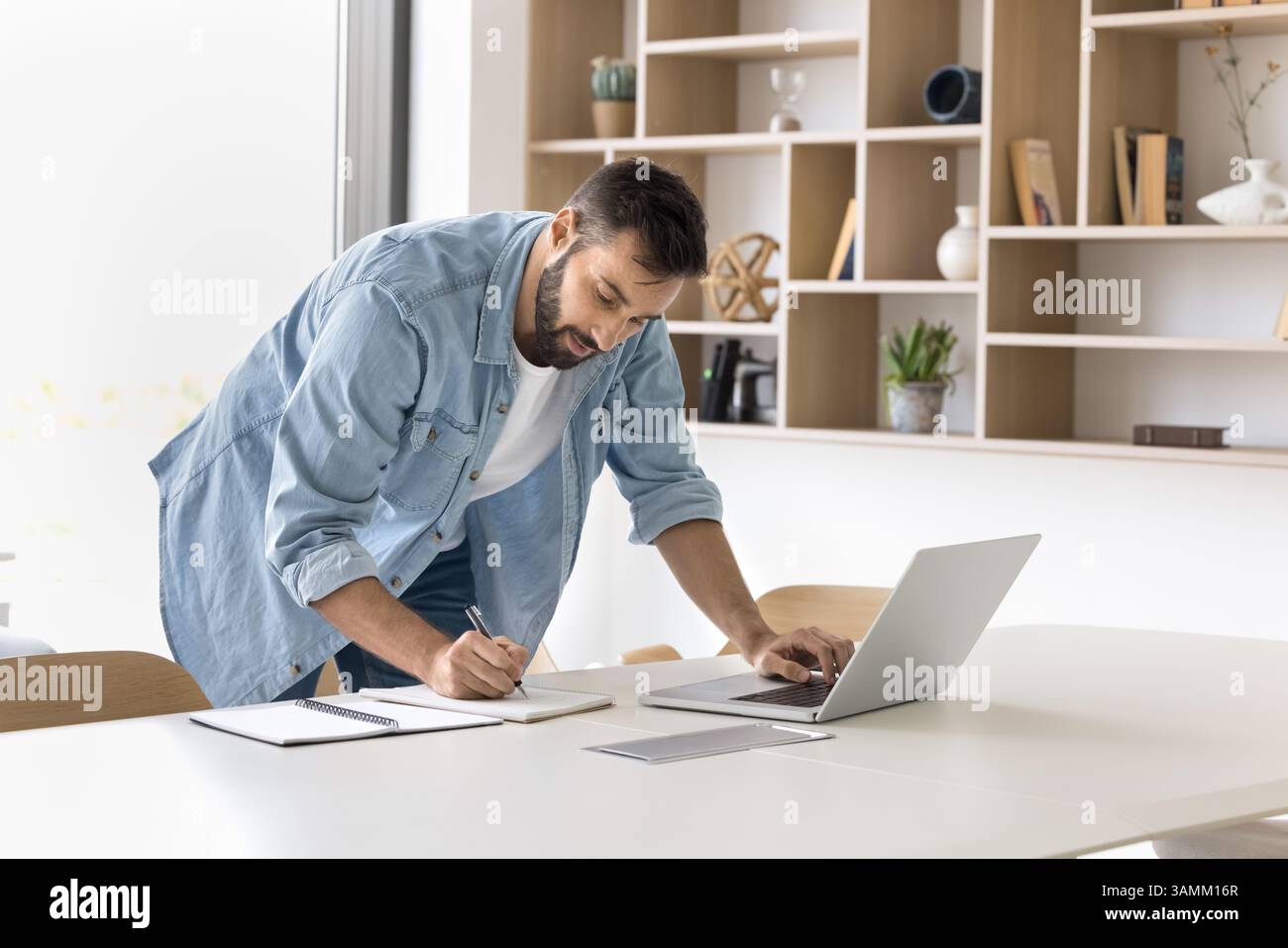 Focused millennial man stand by workplace browse internet write notes Stock Photo
