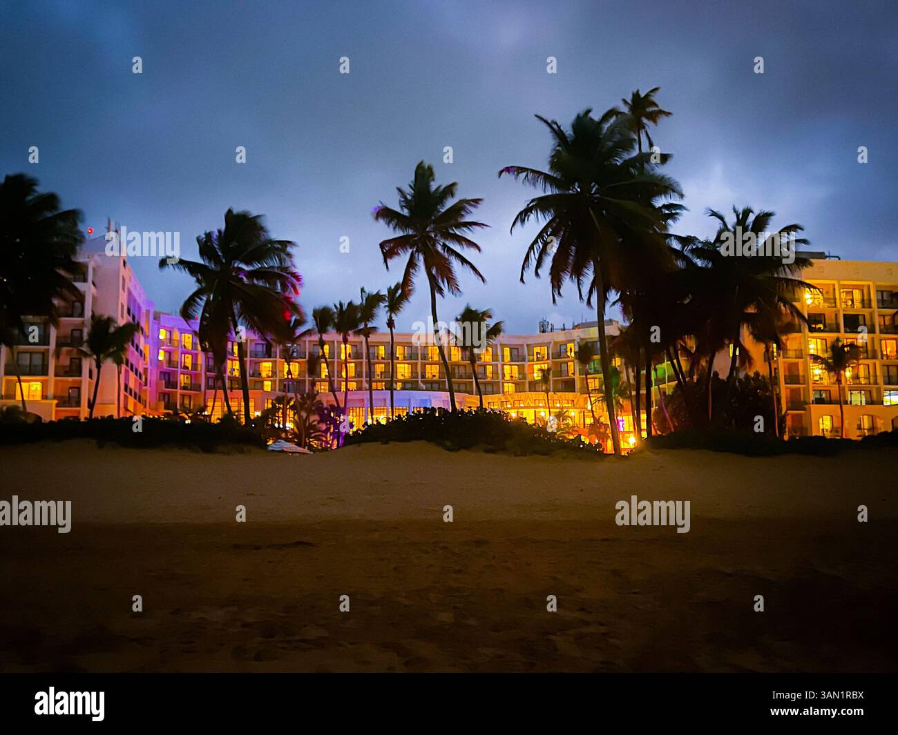 A beachfront resort glows warmly against the backdrop of swaying palms and a moody night sky, capturing the calm before a tropical storm. - Smartphone Captured Stock Image