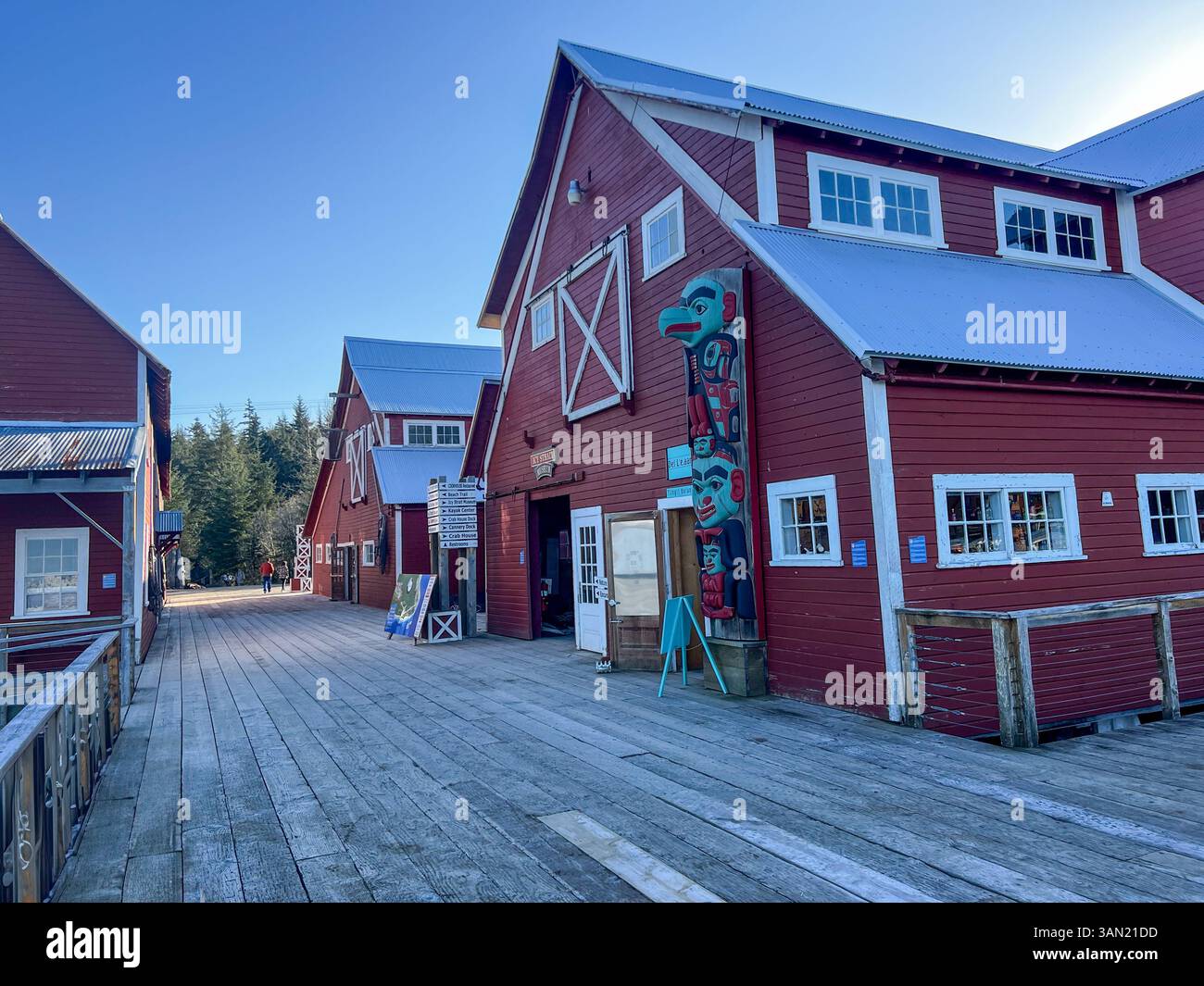 A scenic boardwalk lined with rustic red wooden buildings, featuring a traditional totem pole. A peaceful setting that highlights local culture and hi - Smartphone Captured Stock Image