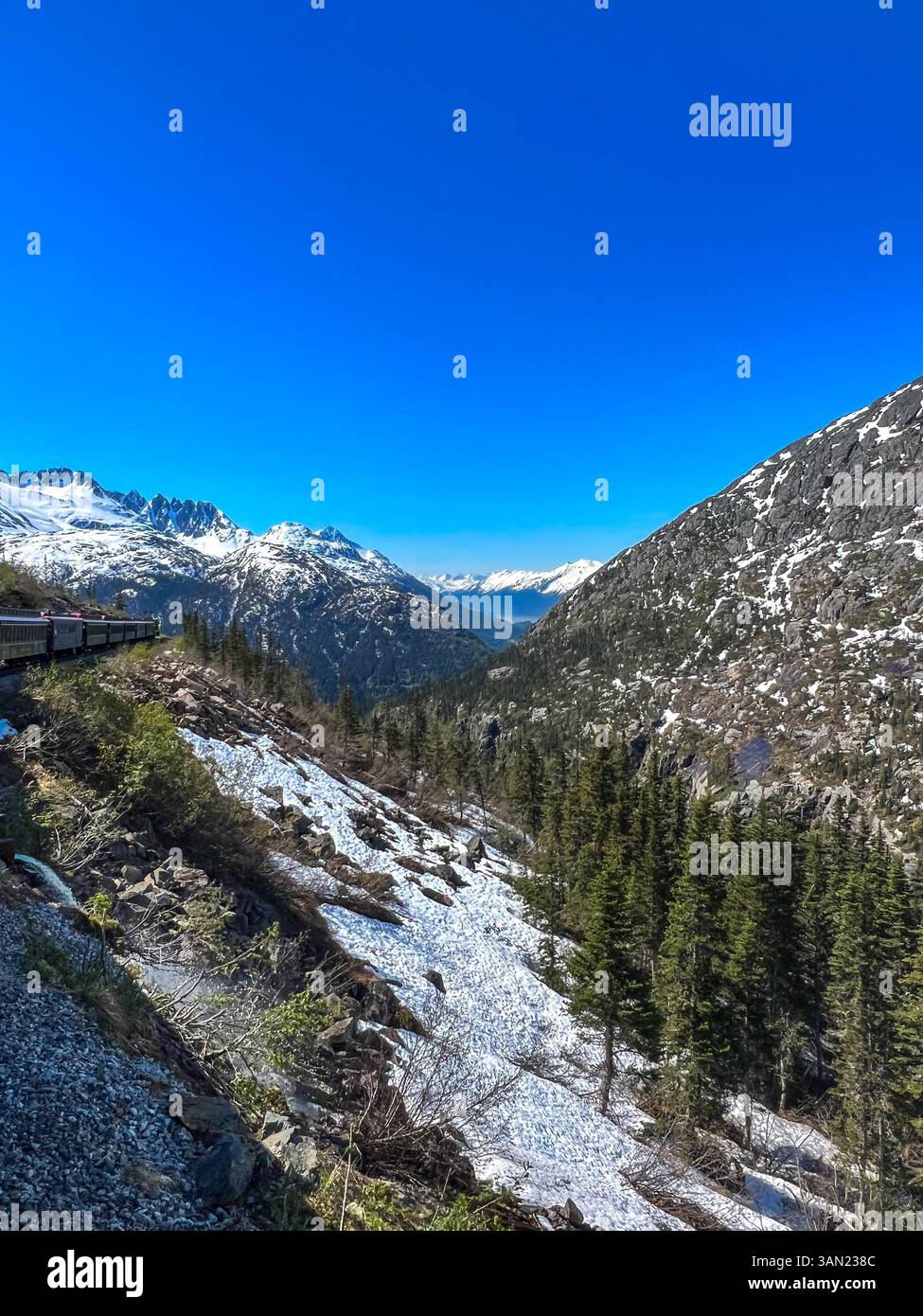 A breathtaking view of a train winding through the rugged Alaskan mountains, passing snow-covered slopes and towering peaks. The crisp blue sky enhanc - Smartphone Captured Stock Image