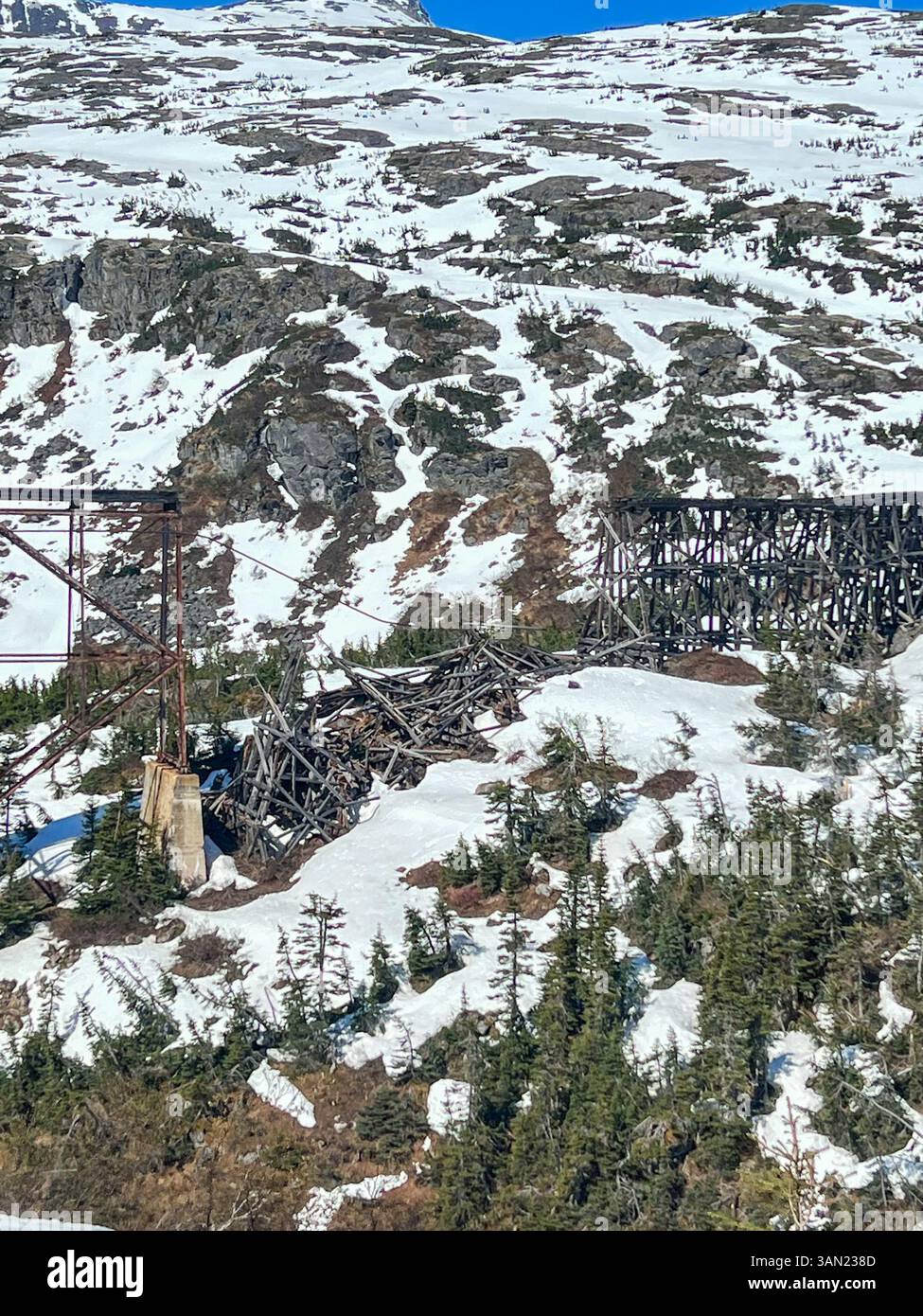 A historic railroad bridge in the Alaskan mountains stands partially collapsed, surrounded by a snow-covered landscape. The remnants of the structure - Smartphone Captured Stock Image