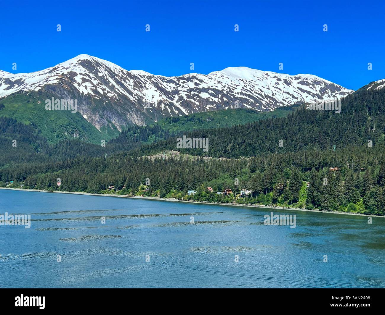 A breathtaking view of Alaska’s snow-capped mountains rising above a dense forest and a tranquil waterfront. Small cabins dot the shoreline, blending - Smartphone Captured Stock Image