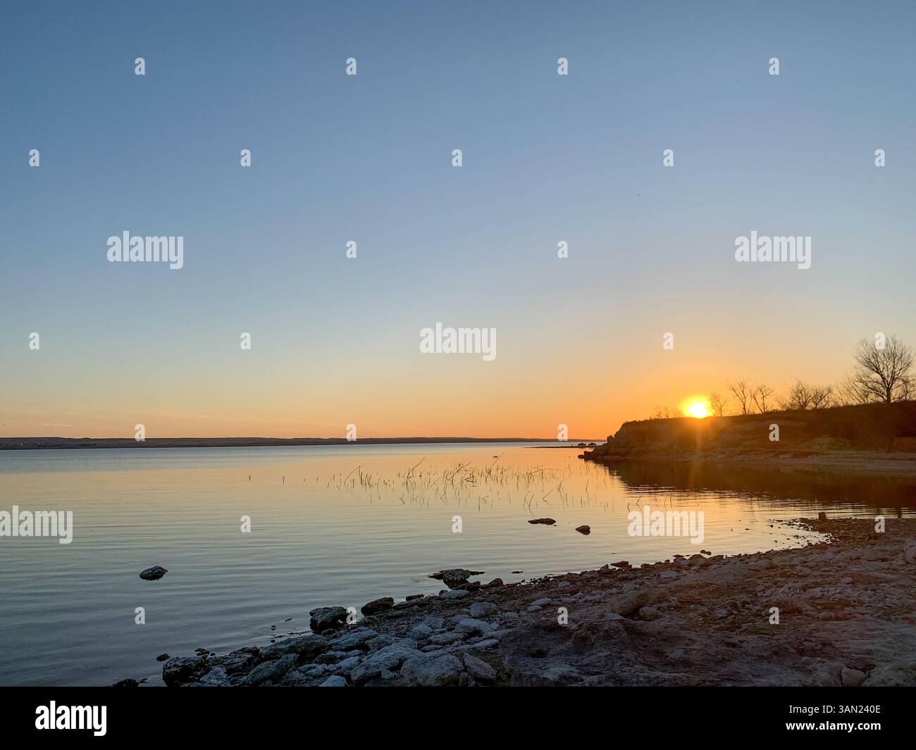 The sun sets over a peaceful lake, casting a golden glow across the calm water. Gentle ripples reflect the fading light, while the rocky shoreline add - Smartphone Captured Stock Image