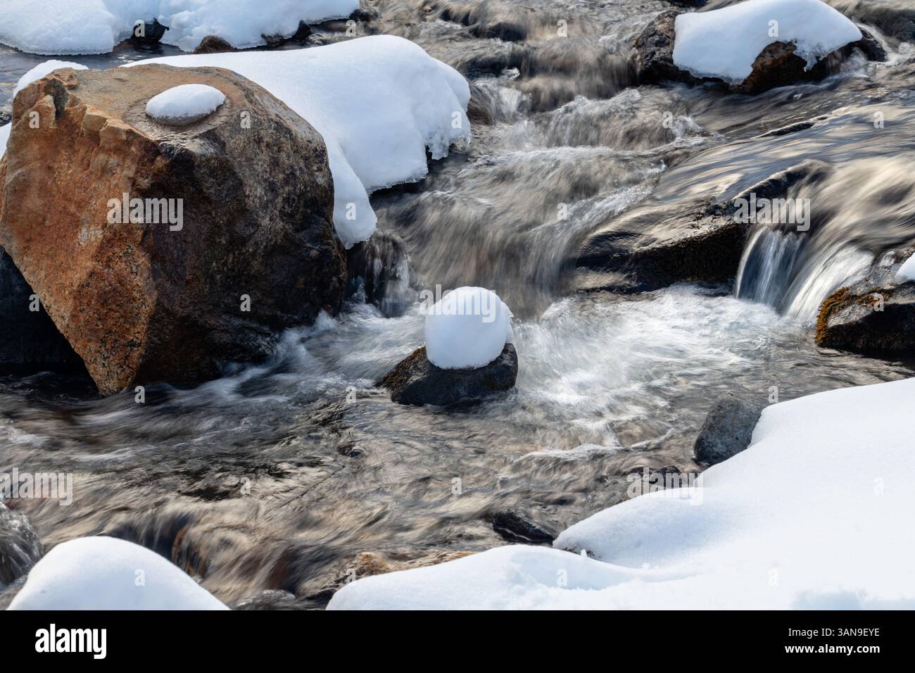A stream of water flows past a large rock covered in snow. The scene is ...