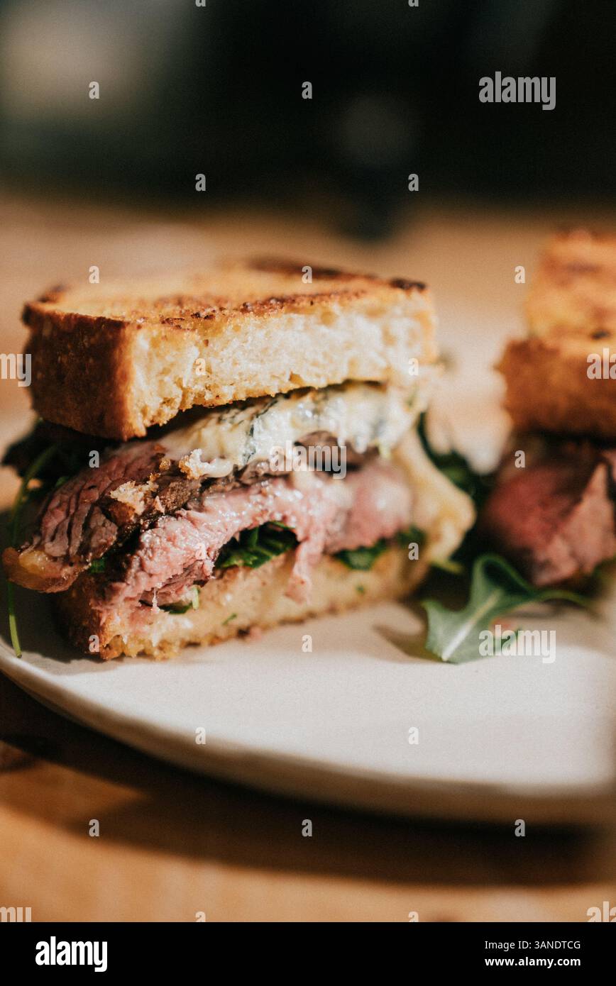 A sandwich with meat and greens is cut in half and placed on a white plate. The sandwich is served on a wooden table Stock Photo