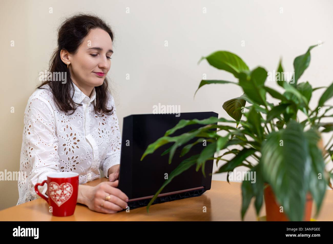 Woman working on laptop with coffee and plant. A young woman in a white blouse sits at a desk using a laptop, with a red coffee mug and a green plant Stock Photo