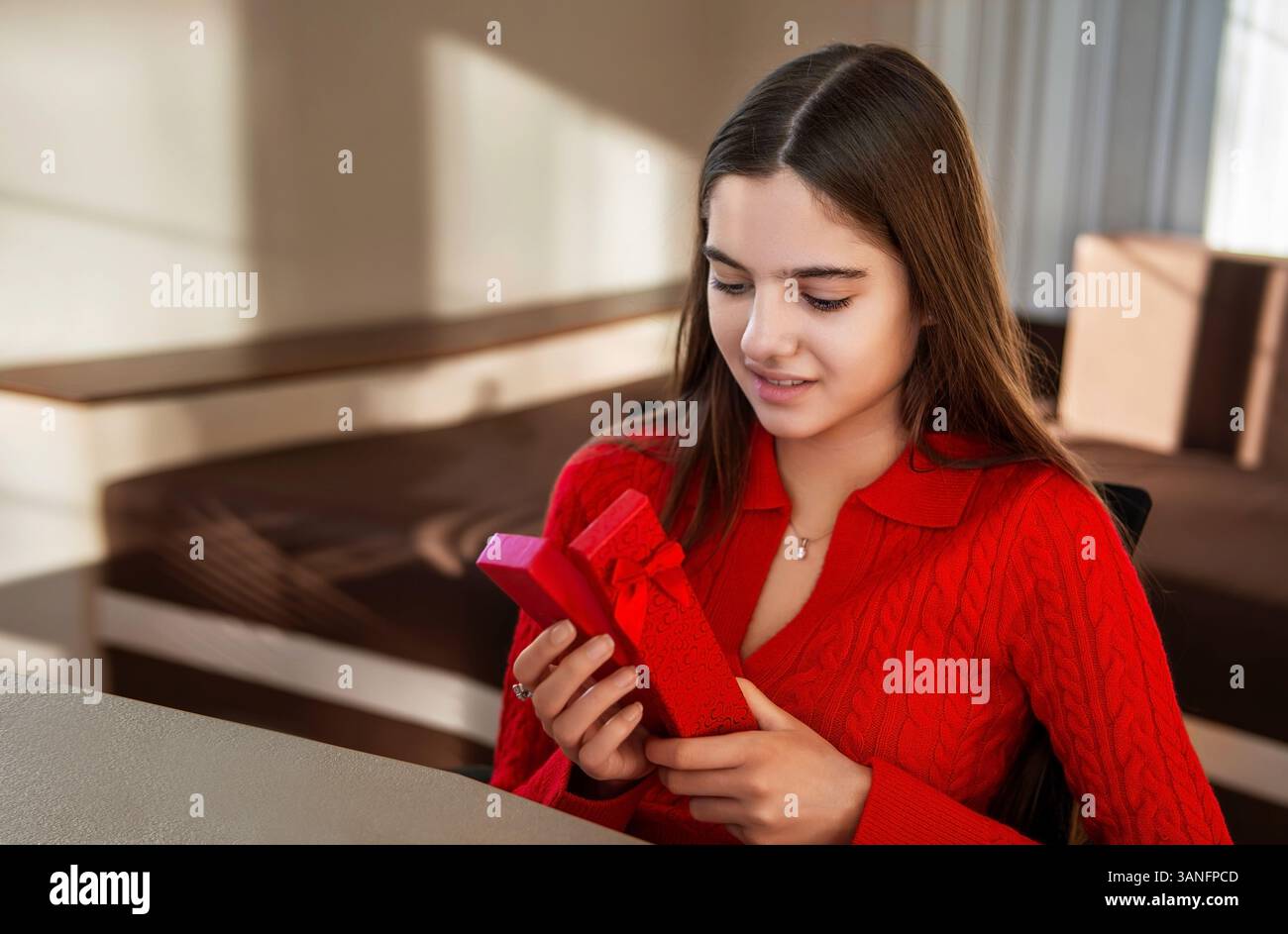 Woman opening a red gift box indoors. A young woman in a red sweater holds and opens a red gift box, expressing curiosity and joy in a cozy room Stock Photo