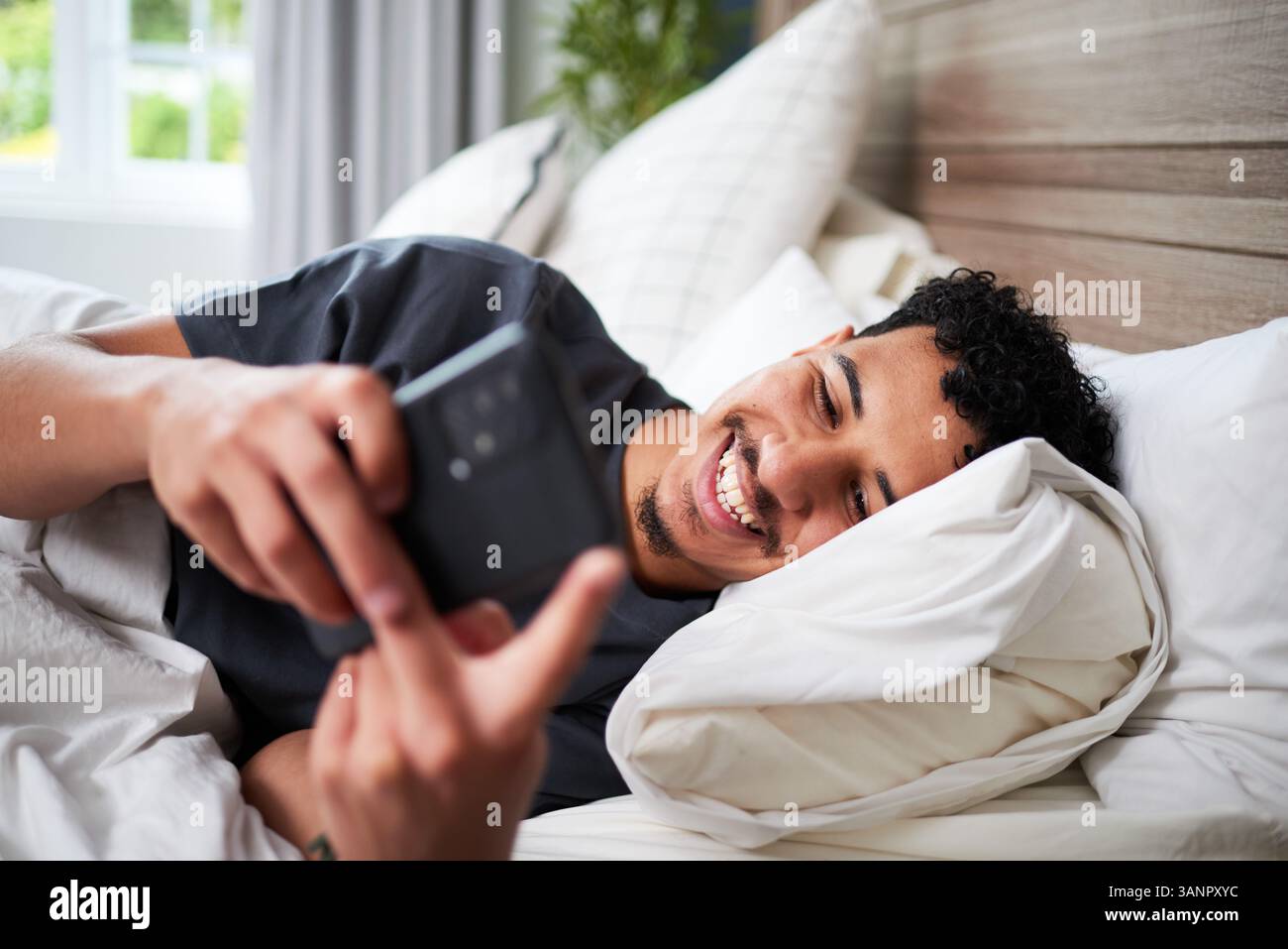 Young Man Relaxing in Bed, Smiling While Using His Smartphone Indoors Stock Photo