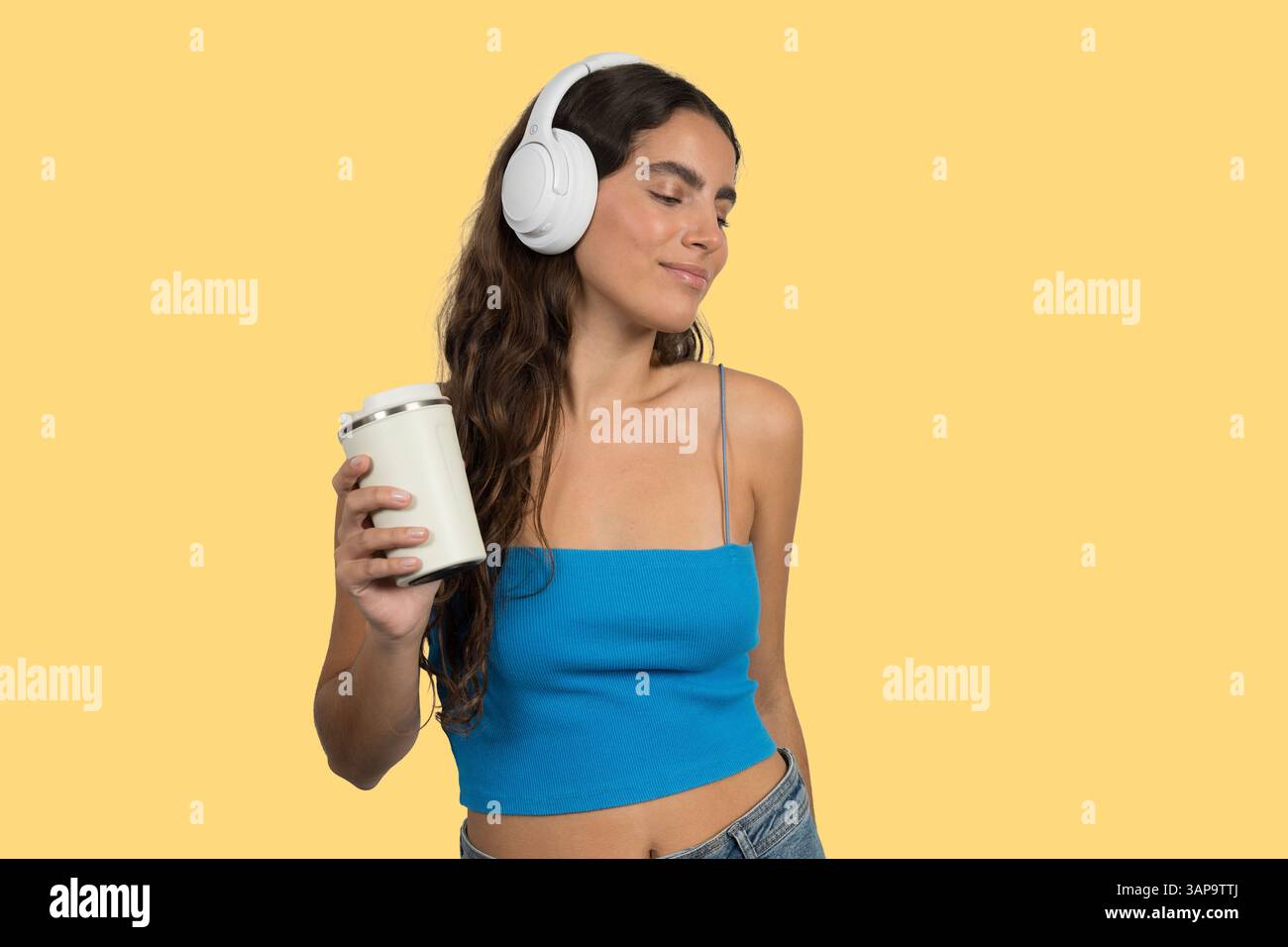 Carefree young woman holding reusable coffee cup while listening music with wireless headphones, isolated on yellow background Stock Photo