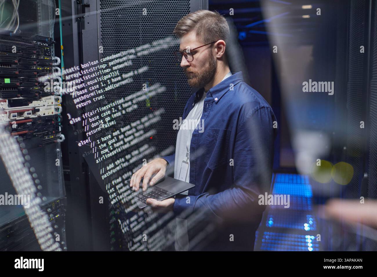 IT professional configuring server settings in modern data center with a laptop. Technical screens displaying code and server racks visible in background Stock Photo