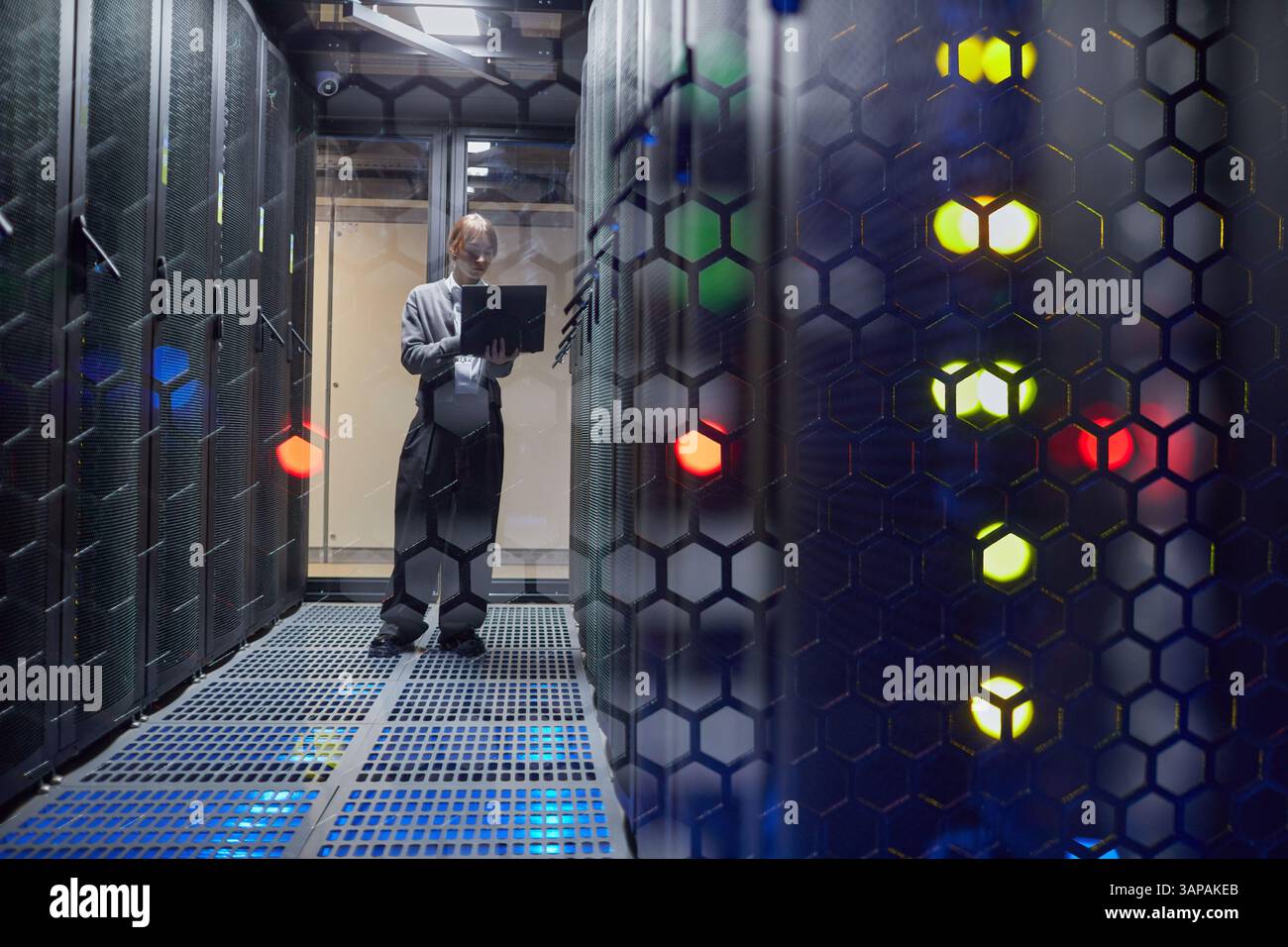 Technician in data center checking system performance on laptop, standing between server racks illuminated by LED indicators Stock Photo
