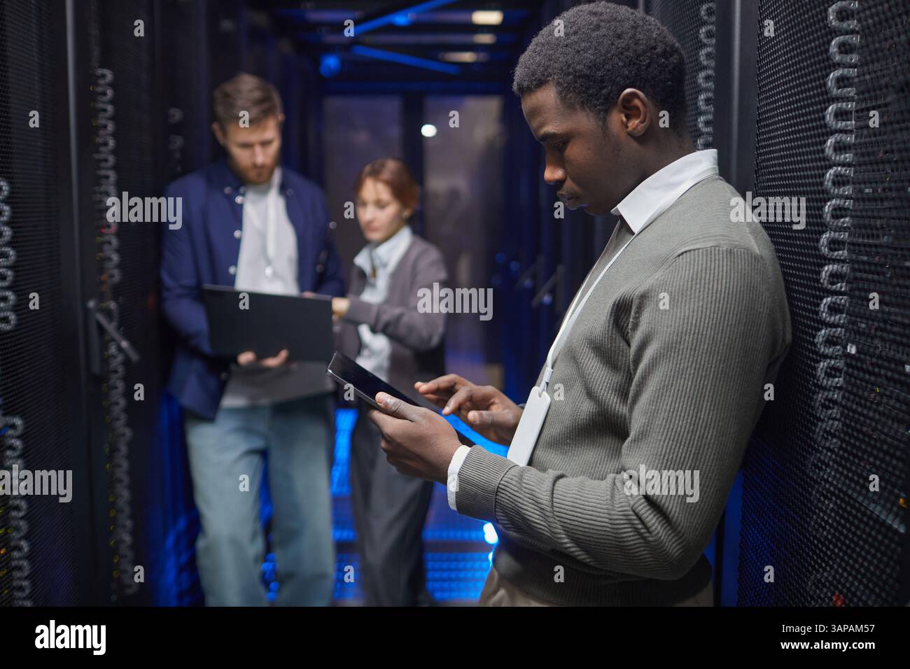 Team of IT professionals working with gadgets in server room analyzing data on devices. Focus on technology and collaboration in high-tech environment Stock Photo