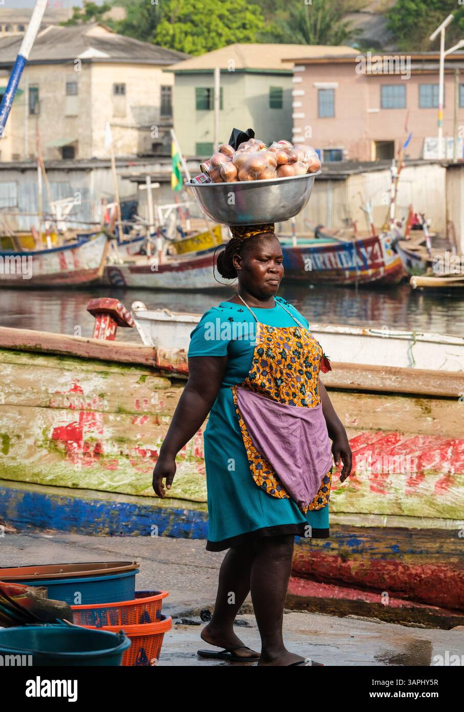 Ghana, Elmina. Woman Walking while Balancing a Basin of Onions on her Head. Stock Photo