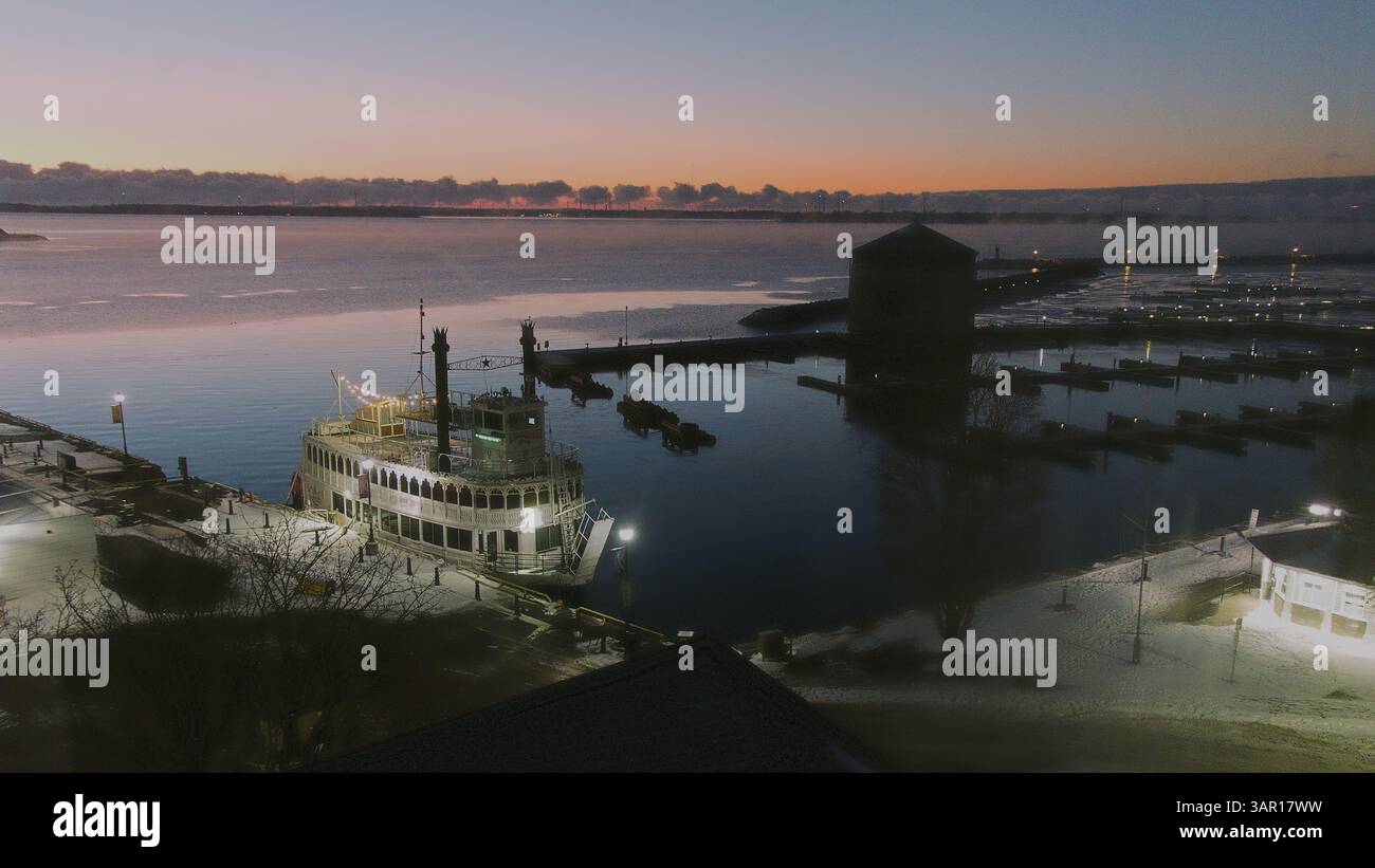 A nostalgic steamboat rests by the icy shore of Kingston, Ontario, as the first hues of dawn rise behind Murney Tower. Lights glow on the deck while t Stock Photo