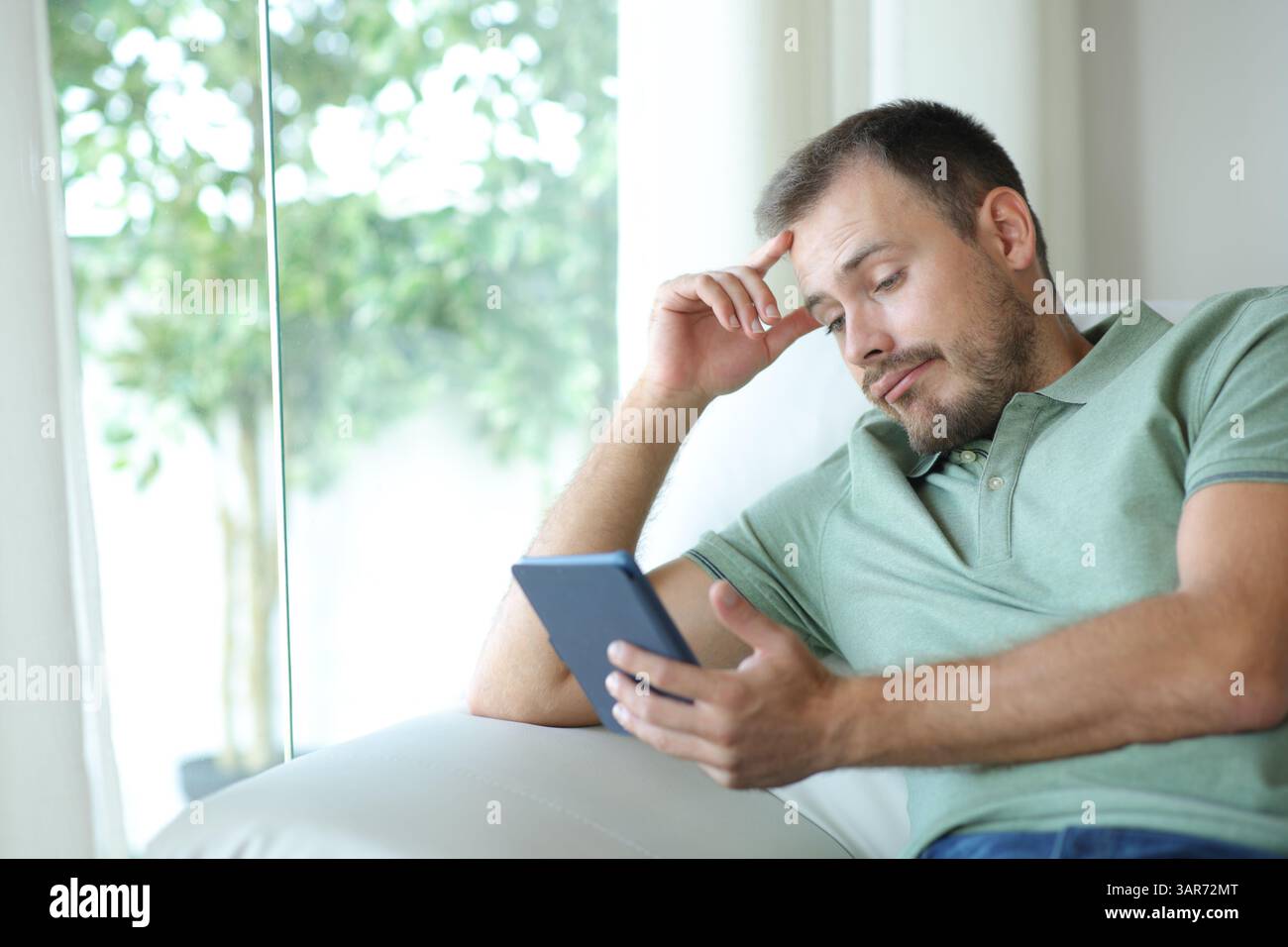 Disappointed man reading a nobel in an ebook sitting on a couch at home Stock Photo