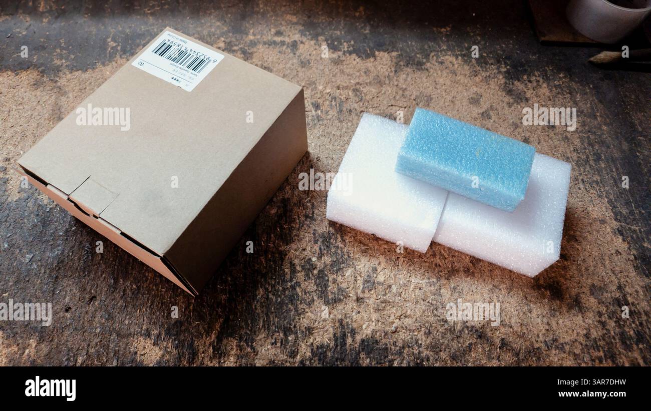 Cardboard shipping box and protective foam padding materials sit on a dusty workbench, suggesting packing or unpacking activities in a workshop enviro Stock Photo