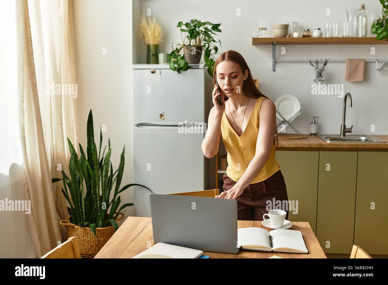 A young woman talks on the phone while getting ready to work at her kitchen table. Stock Photo
