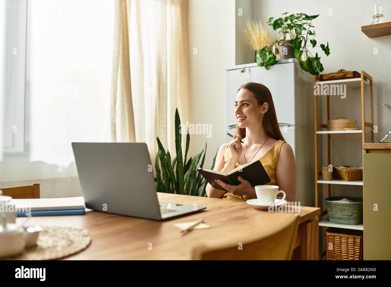 Beautiful woman smiles while noting ideas in her notebook at a bright wooden table. Stock Photo