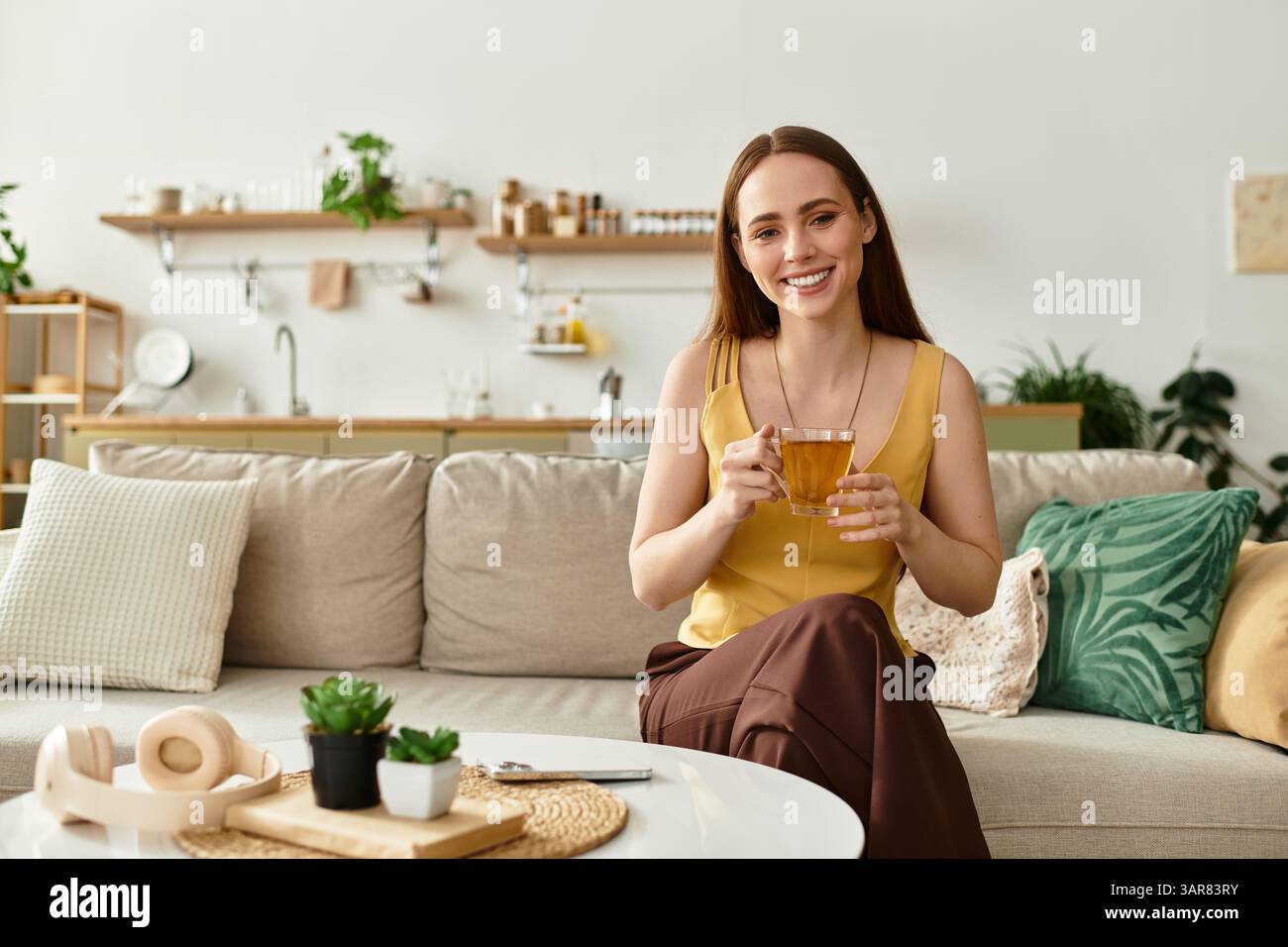 Joyful woman sips tea on the couch, surrounded by greenery and cozy decor. Stock Photo