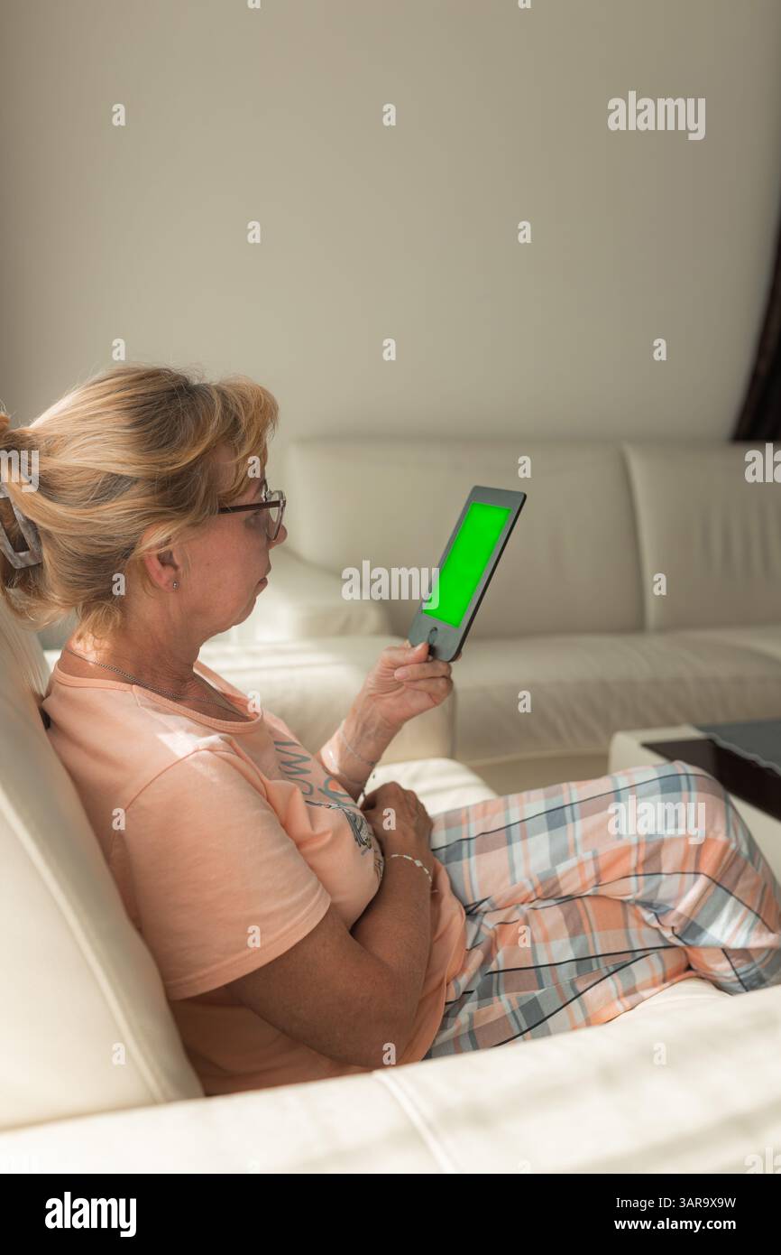 Senior woman using e-reader with green screen on couch Stock Photo