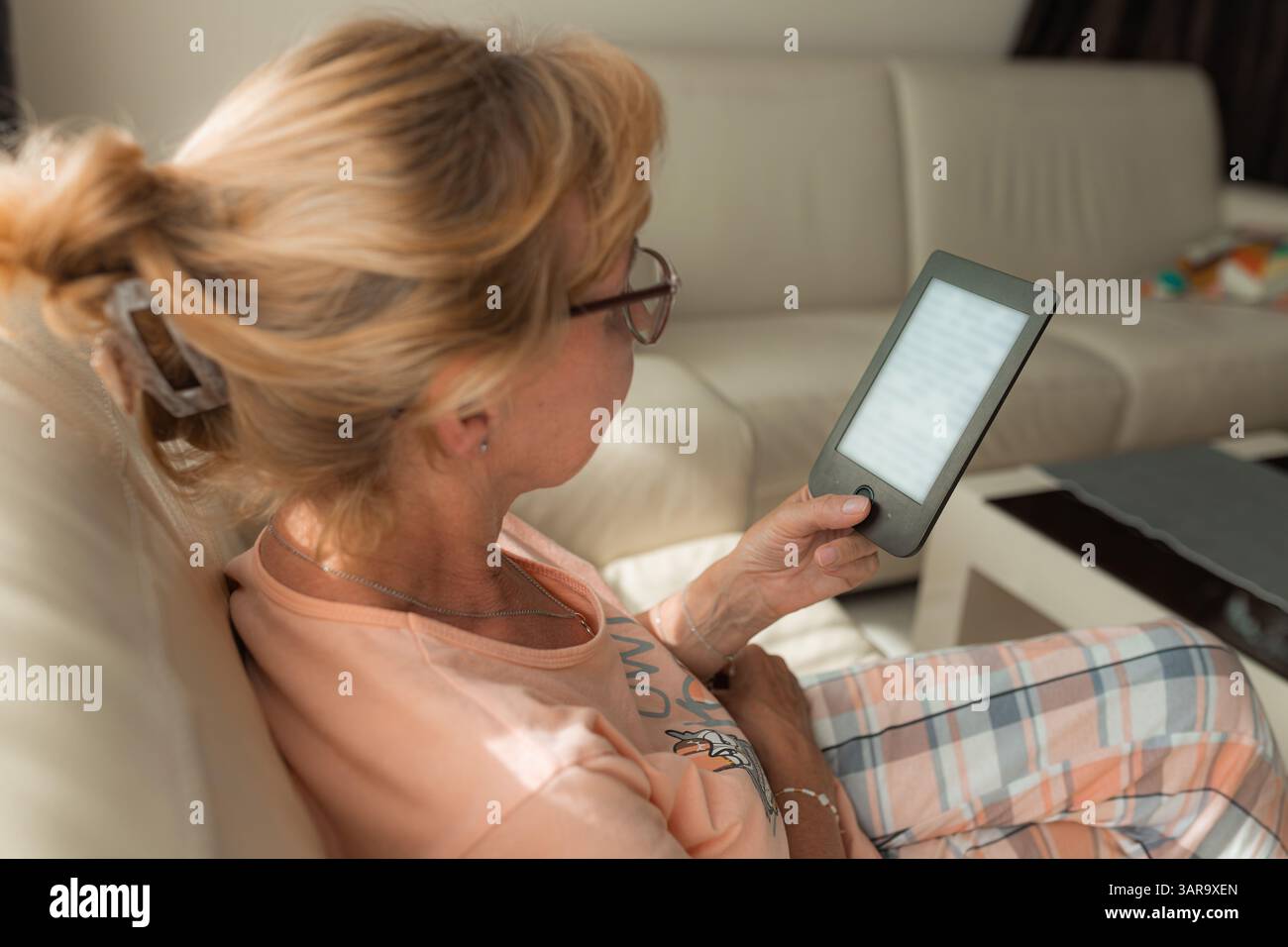 Side view of a mature woman reading an e-book on her e-reader, sitting on white couch in cozy home environment. Stock Photo