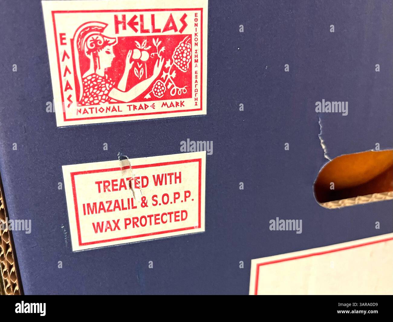 Hellas cardboard crate of oranges featuring chemical warning label indicating treatment with imazalil and S.O.P.P. wax for post-harvest protection. - Smartphone Captured Stock Image