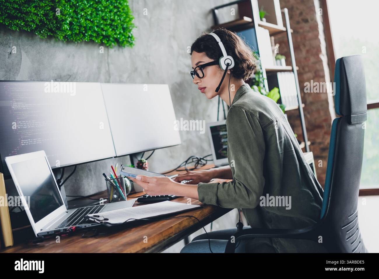 Young female programmer working remotely in a stylish home office with dual monitors, coding software and managing tasks efficiently Stock Photo
