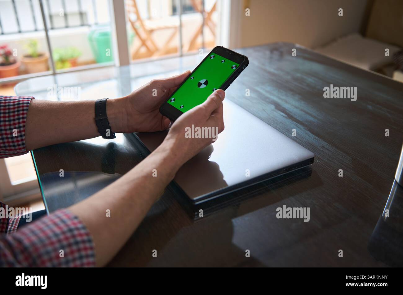 Man interacts with a smartphone displaying a green screen, seated at a table indoors. Nearby, a sleek laptop rests on the desk, depicting a workspace Stock Photo