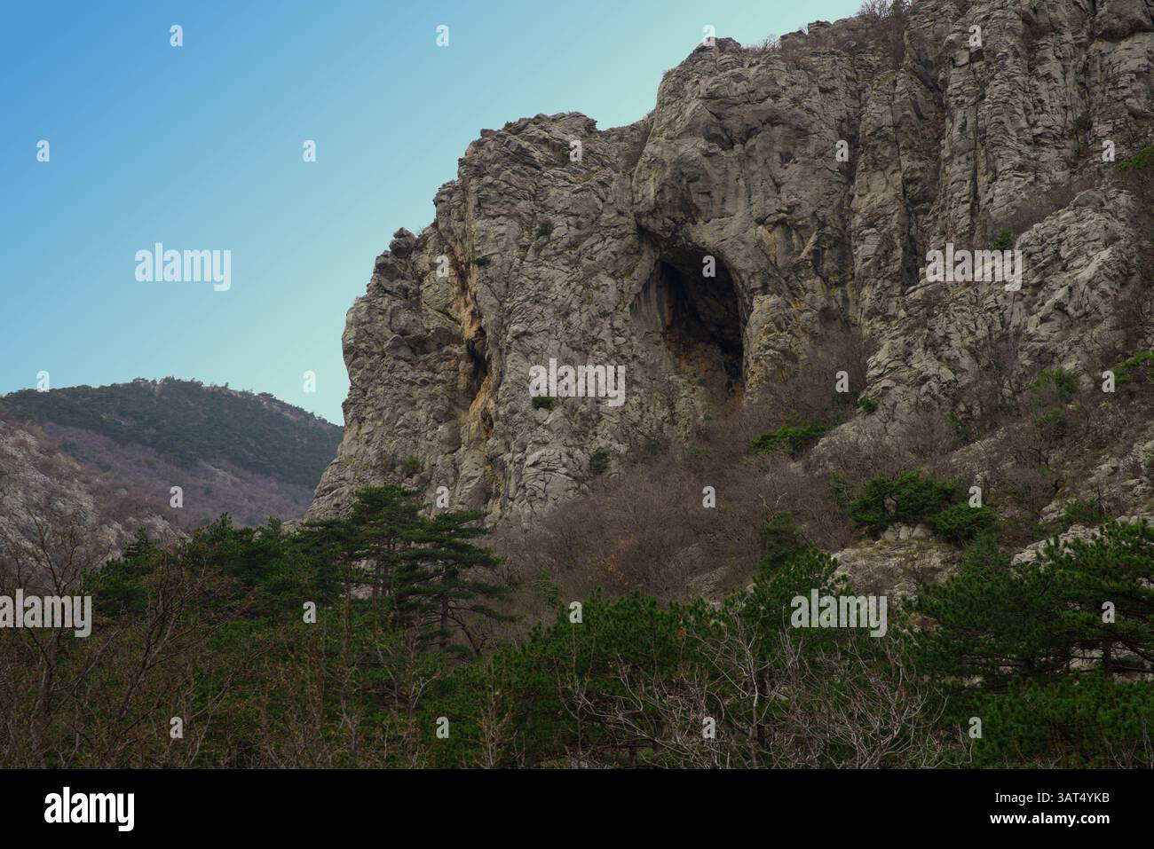 The image captures a rugged mountain landscape with a prominent rocky cliff and large cave, surrounded by dense vegetation under a clear blue sky Stock Photo