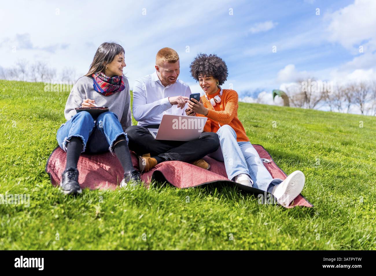 Three students are sitting on grass using laptop and smartphone, enjoying studying together in a park Stock Photo
