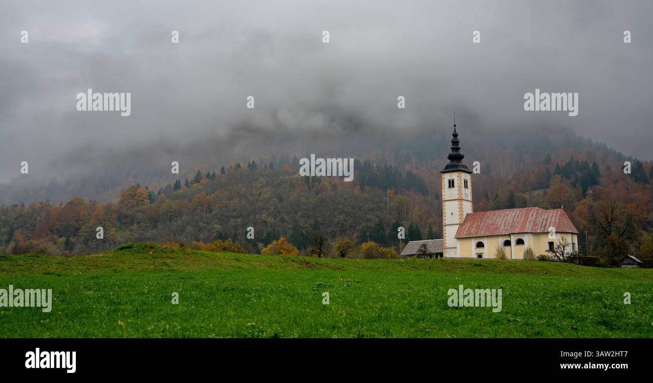 Low clouds Cerkev Marijinega church at Bitnje nestled in a colorful autumn forest, creating a serene and picturesque landscape. Slovenia countryside Stock Photo