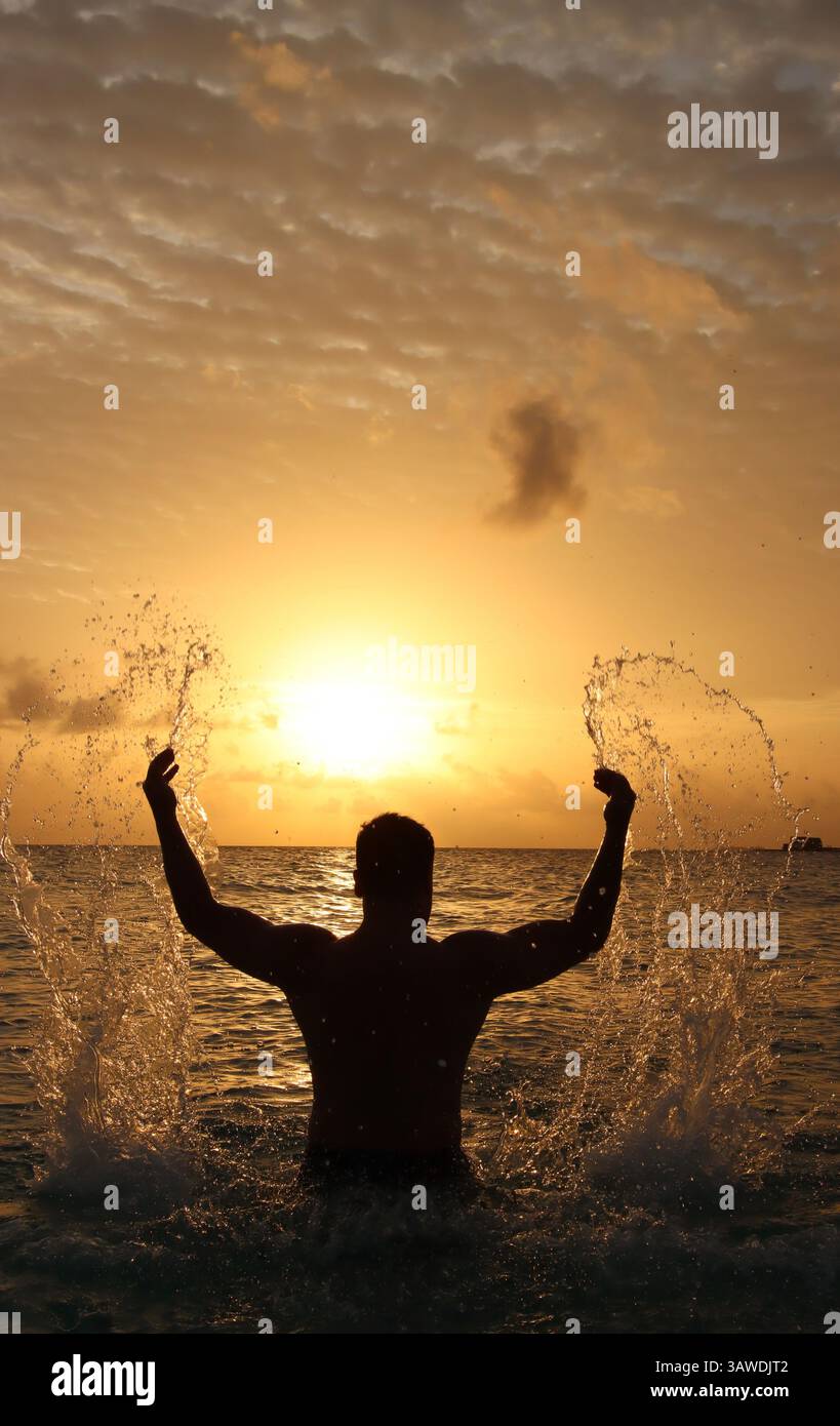 Silhouette of a man playfully splashing water as the sun sets over the ocean, creating a stunning backdrop of warm colors and dramatic clouds. Stock Photo