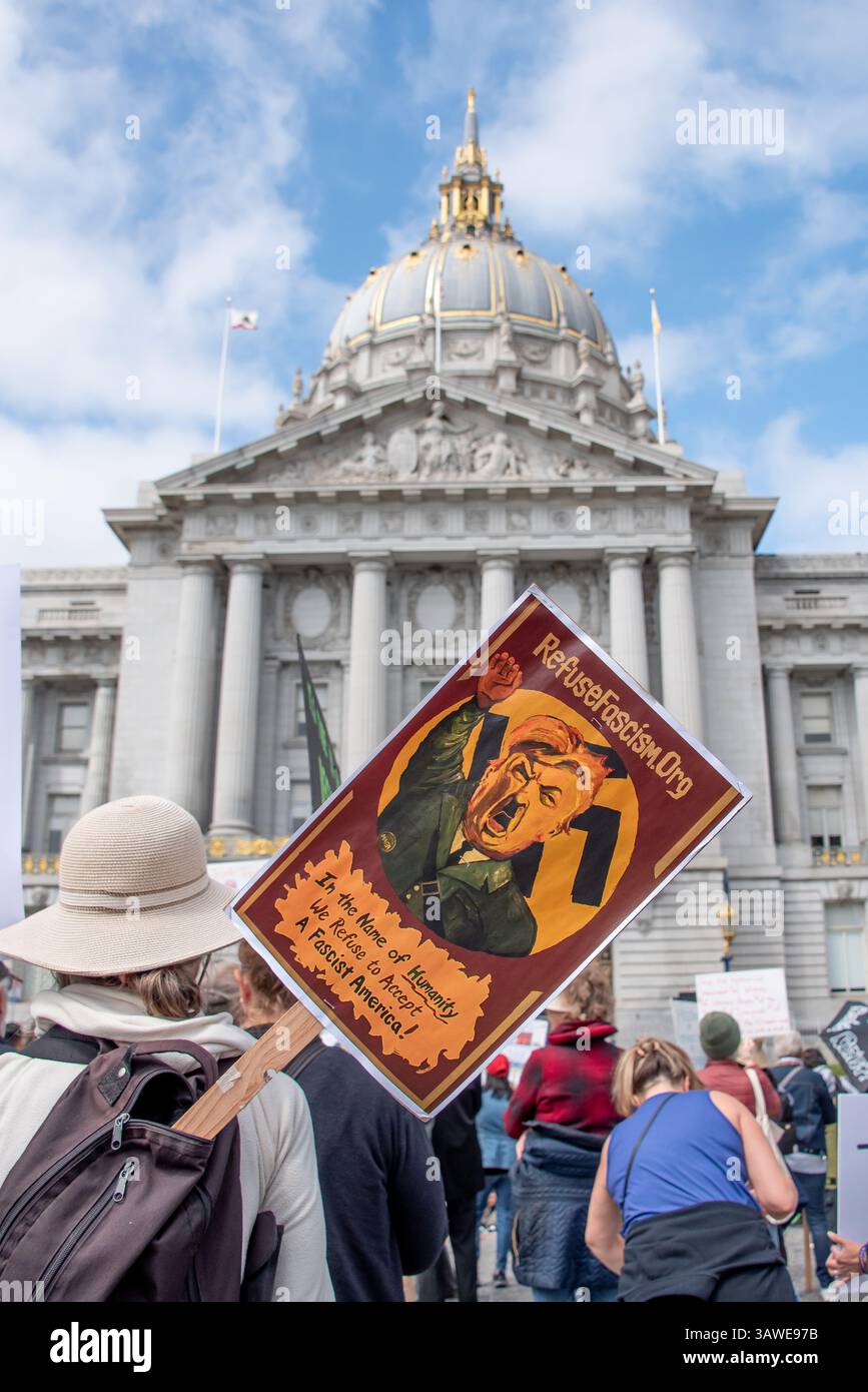 San Francisco, USA. 19th Apr 2025. At Civic Center Plaza, in front of San Francisco City Hall, protesters gather for the “Stop the Billionaire Agenda” protest against the Trump Administration. A protest sign sticks out from a protester's backpack reading, 'In the name of Humanity, We Refuse to Accept a Fascist America!' A retro-looking illustration of Trump as Hitler giving a Nazi salute appears above the text. Credit: Shelly Rivoli/Alamy Live News - Stock Image