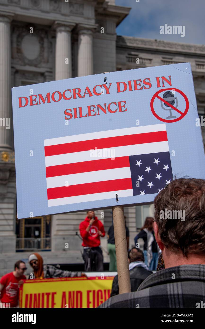 San Francisco, USA. 19th Apr 2025. At Civic Center Plaza during the “Stop the Billionaire Agenda” protest against the Trump Administration., a man listens to speakers while holding a sign reading, 'Democracy dies in silence,' showing an upside down American flag and a microphone with a red line through it. Credit: Shelly Rivoli/Alamy Live News - Stock Image