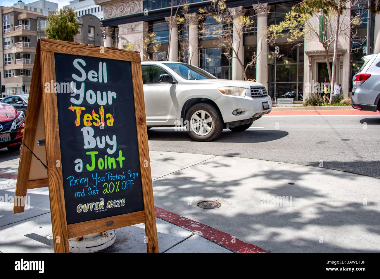 San Francisco, USA. 19th Apr 2025. While protesters gather outside the Tesla showroom and dealership in San Francisco, the Outer Haze cannabis shop across the street places a humorous sign on the sidewalk reading, 'Sell Your Tesla, Buy a Joint.' and telling protesters, 'Bring your protest sign and get 20% off.'  Credit: Shelly Rivoli/Alamy Live News - Stock Image