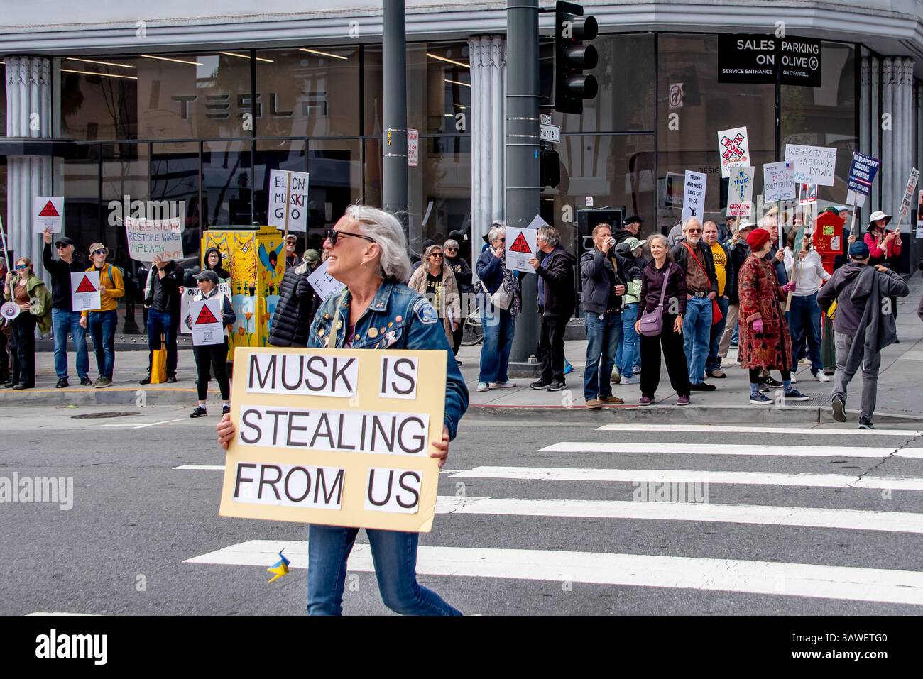 San Francisco, USA. 19th Apr 2025. Protesters gather outside the Tesla showroom and dealership in San Francisco, shown in the background as a woman rosses the street in front of them carrying a sign reading, 'Musk is stealing from us.'  Credit: Shelly Rivoli/Alamy Live News - Stock Image
