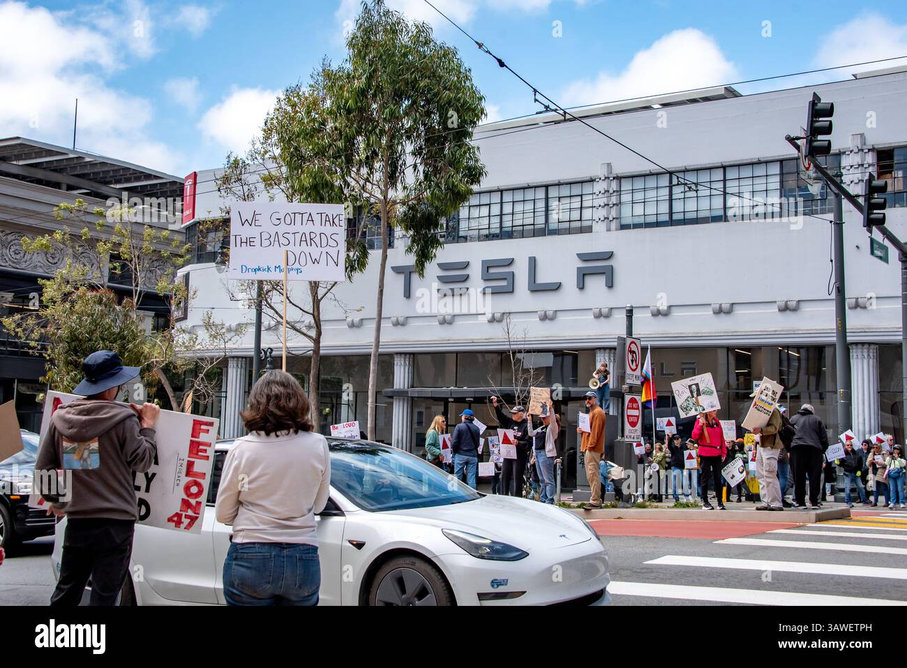 San Francisco, USA. 19th Apr 2025. Protesters gather outside the Tesla showroom and dealership in San Francisco and on all sides, including the islands between street lanes, at the intersection of O'Farrel and Van Ness. A white Tesla car drives through as signs can be read saying, 'Felon 47,' and 'We gotta take the bastards down.'  Credit: Shelly Rivoli/Alamy Live News - Stock Image