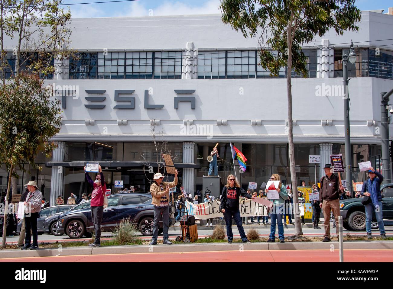 San Francisco, USA. 19th Apr 2025. Protesters gather outside the Tesla showroom and dealership in San Francisco, some along the island running down the middle of Van Ness Avenue. BEhind them waves a rainbow flag and a young woman stands high atop a utility box speaking into a megaphone.  Credit: Shelly Rivoli/Alamy Live Newsa - Stock Image