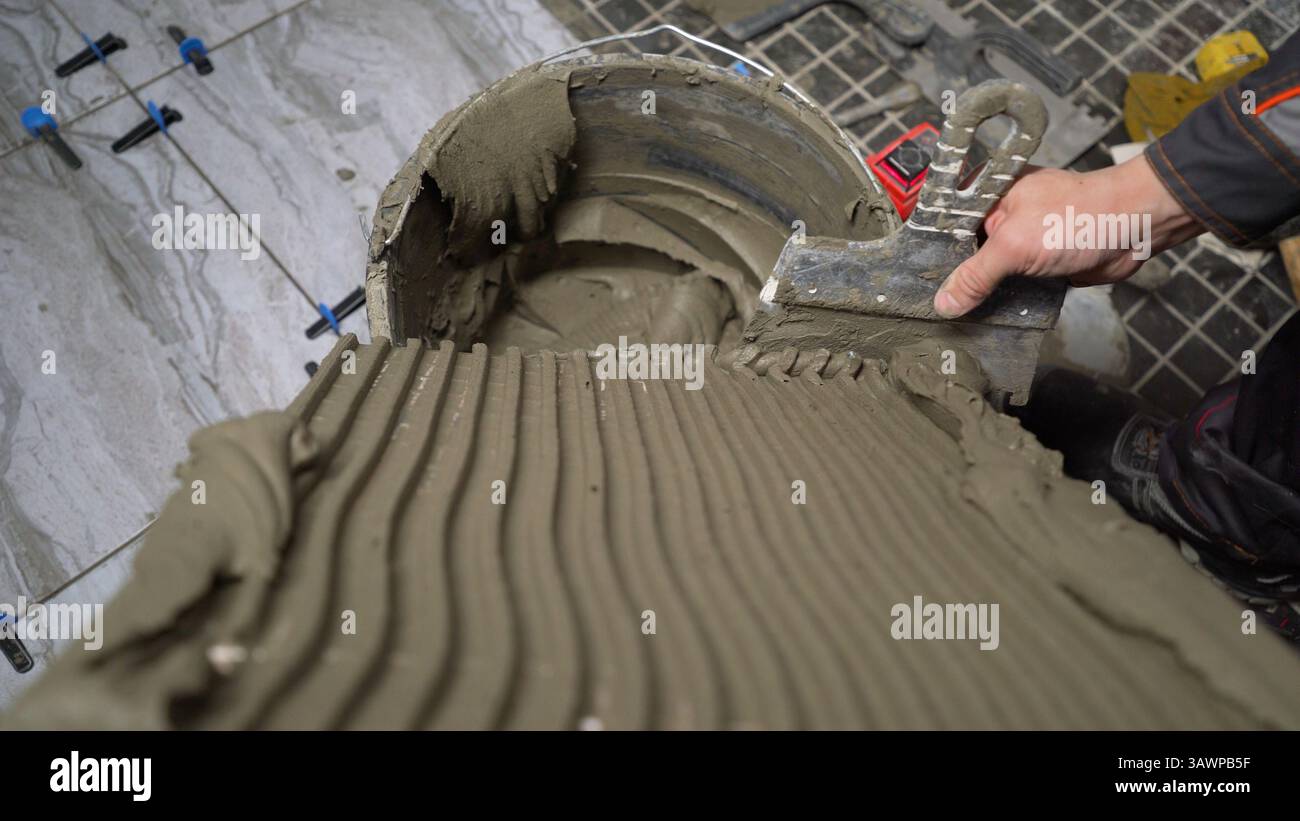 Construction worker spreading tile adhesive on floor using notched trowel, preparing for tile installation Stock Photo