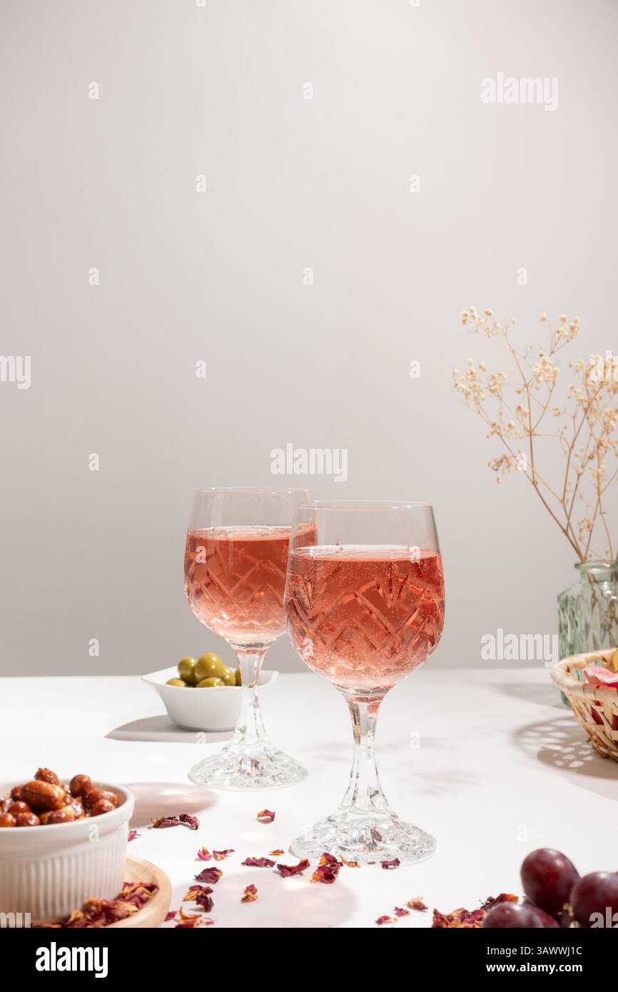 Two glasses of rose wine on a white tablecloth. On the table there are a small bowl with peanuts, red grapes, a bread basket and a green vase. Stock Photo