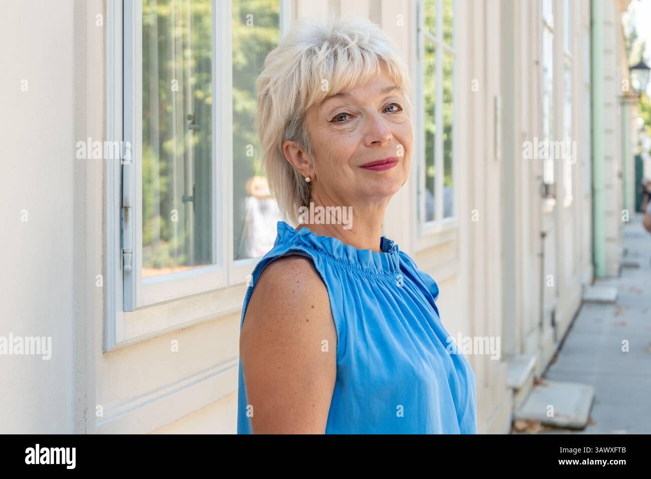 A stylish woman aged 65-70 in a blue outfit stands outside a building with window. Stock Photo