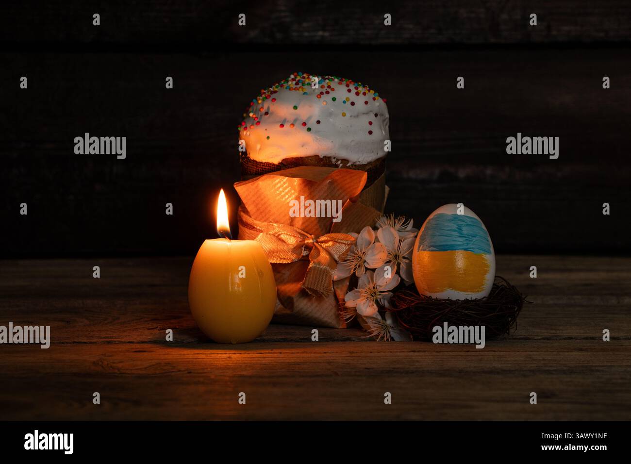Easter egg with painted flag of Ukraine, kulich and blossoming branch of cherry and candle in the dark on wooden table, Easter in Ukraine Stock Photo