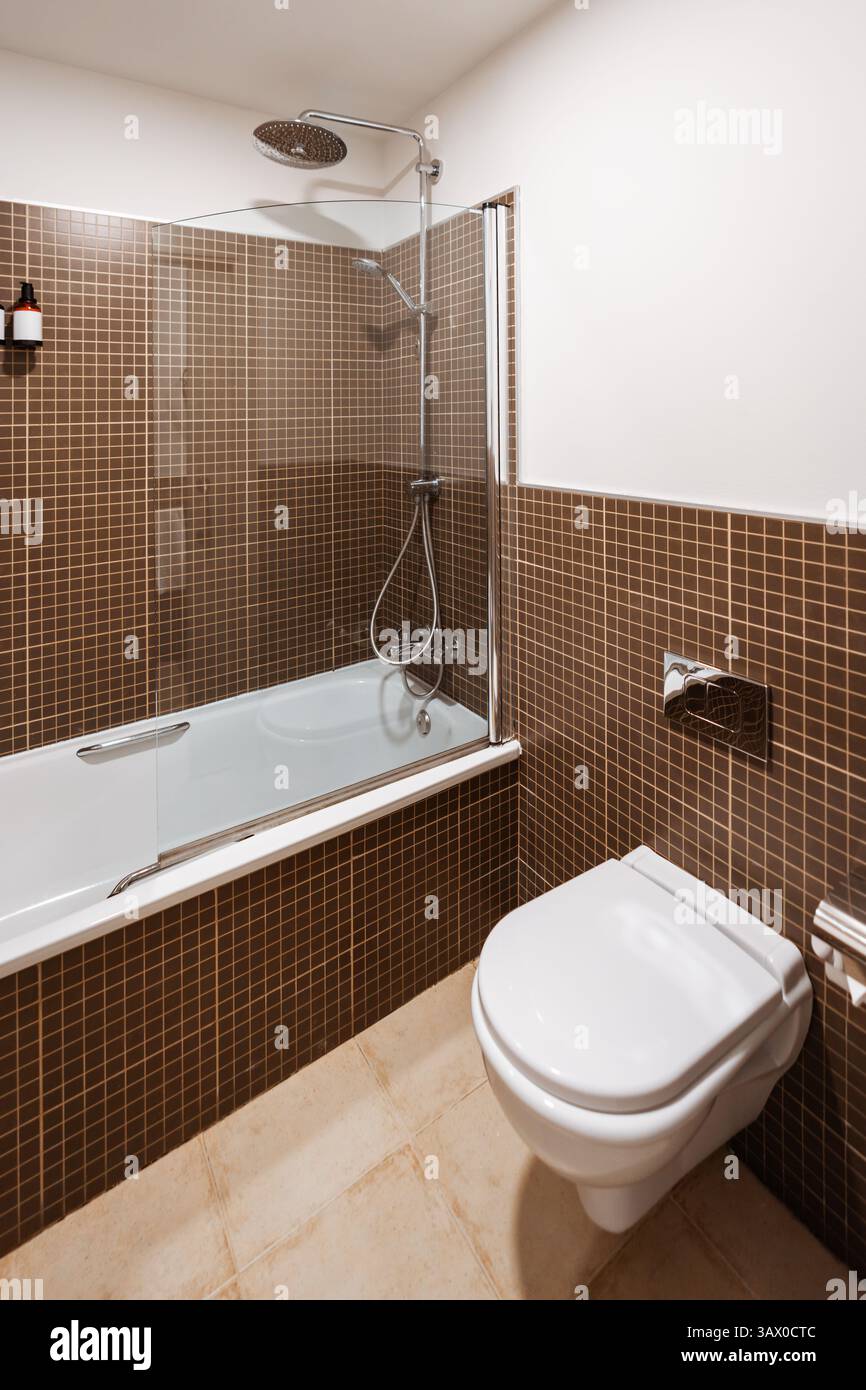 Modern bathroom with wall-mounted toilet, white bathtub with overhead rain shower and handheld shower, brown mosaic wall tiles, glass partition, ceram Stock Photo