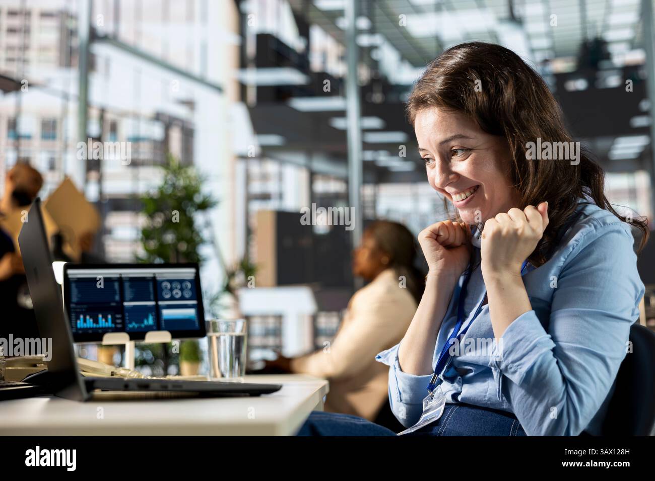 Happy business worker celebrating a major project milestone at her desk, thrilled with the successful outcome. Enthusiastic woman smiles and enjoys good news, satisfied of professional victory. Stock Photo